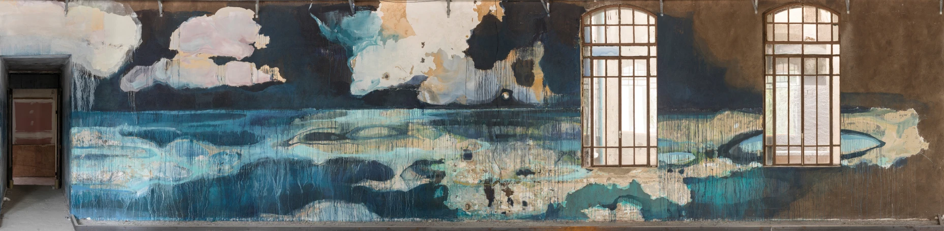 Parede interna de um prédio antigo com um grande mural pintado em tons de azul, verde e bege, evocando uma paisagem aquática com nuvens no céu. A pintura aparece desgastada, com escorridos de tinta. Duas janelas altas com molduras metálicas interrompem o mural, deixando a luz entrar. À esquerda, uma porta escura contrasta com as cores da obra.
